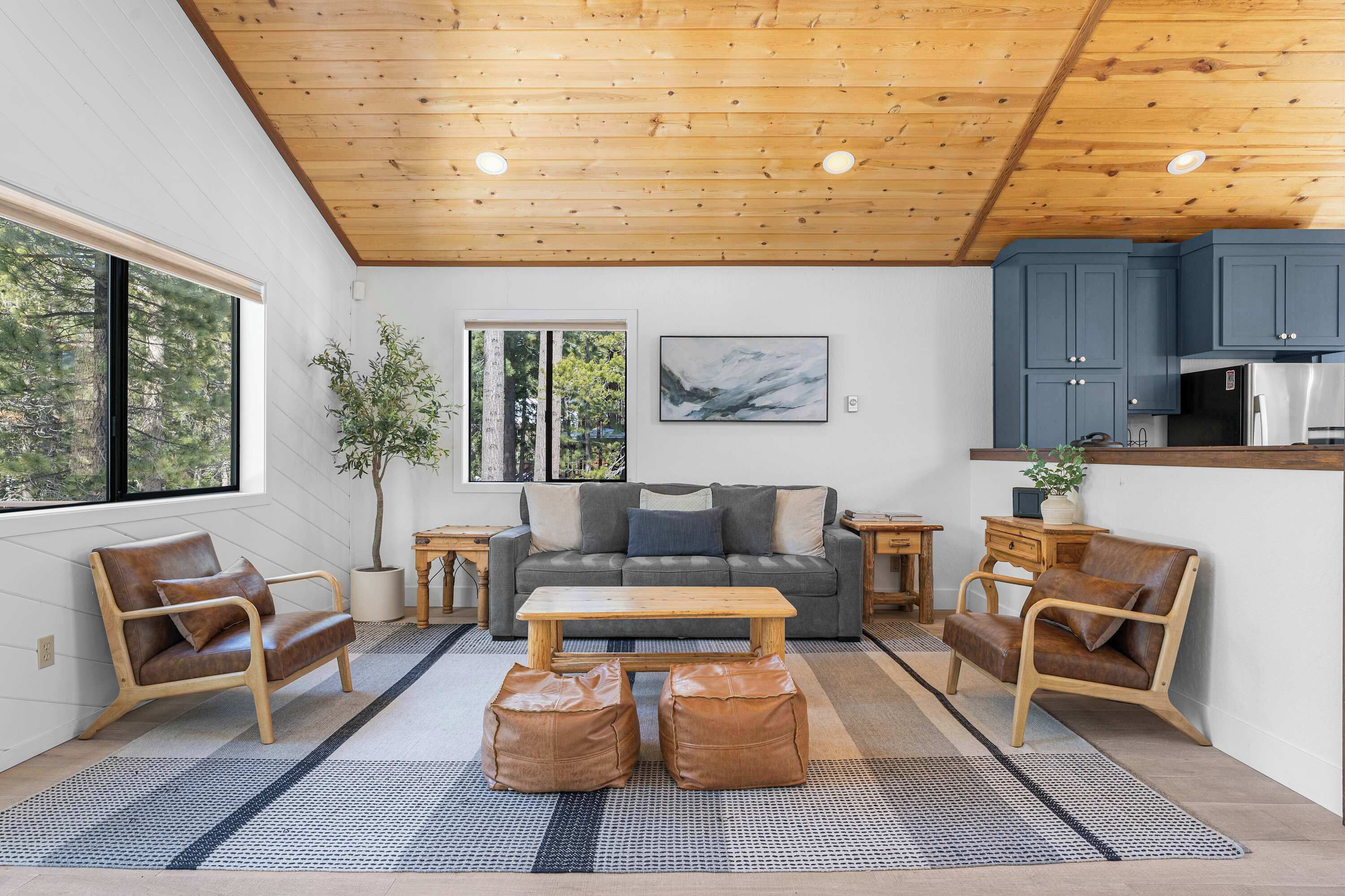 Un salon moderne avec un plafond en bois, un canapé gris, des fauteuils en cuir, une table basse en bois et un coin cuisine avec des meubles bleus.