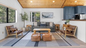 Una sala de estar moderna con techo de madera, un sofá gris, sillones de cuero, una mesa de centro de madera y una zona de cocina con armarios azules.
