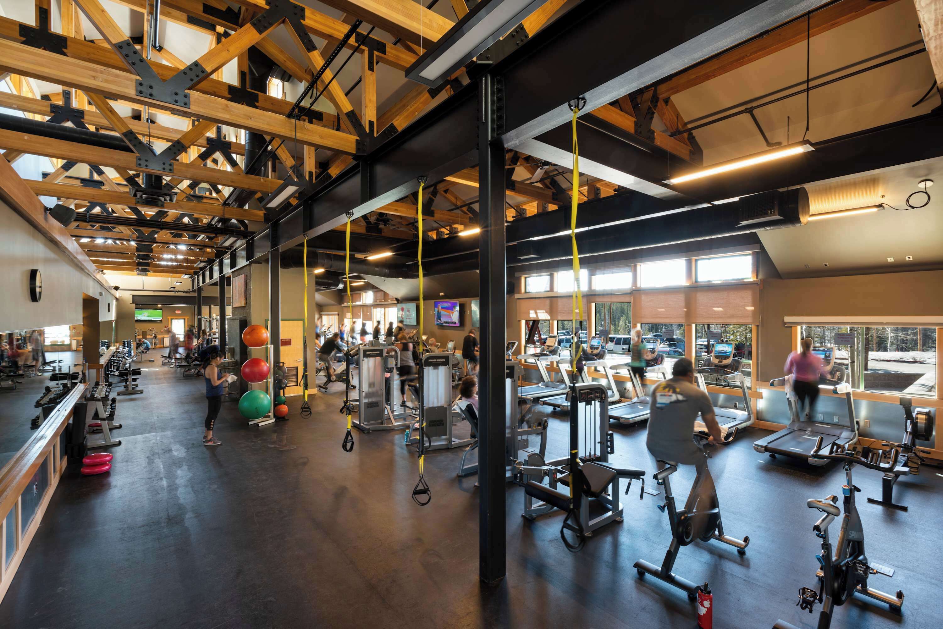 Une salle de sport équipée de divers appareils d’exercice, avec des poutres en bois au plafond et de grandes fenêtres.