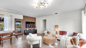 A modern living room with a wooden floor, a round coffee table, a white sofa, and a dining area with wooden chairs. - 4BR Biscayne River Home with Pool & Pickleball Fun (North Miami Beach)