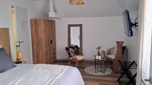 Un dormitorio con una cama, un armario de madera, un espejo, sillas de mimbre, una mesita con objetos y un televisor montado en la pared.