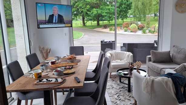 Un comedor con una larga mesa de madera dispuesta para una comida, una pantalla de televisión mostrando un noticiero y vistas de un jardín ajardinado a través de grandes ventanales.