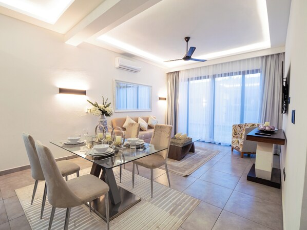 A modern living room with a glass dining table, comfortable seating, a ceiling fan, and large windows with sheer curtains.