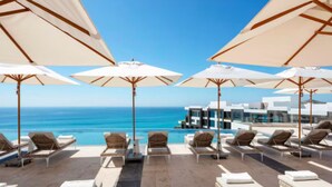 A poolside area with lounge chairs and umbrellas, overlooking a clear blue sea and a modern building. - Tranquil 1 Bedroom Suite in Cabo San Lucas (Cabo San Lucas)