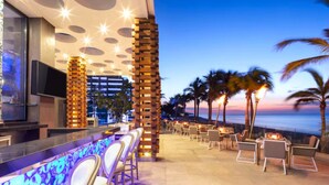 An outdoor bar area with a view of the ocean at dusk, featuring a modern design with a bar counter, seating, and a decorative pillar. - Tranquil 1 Bedroom Suite in Cabo San Lucas (Cabo San Lucas)