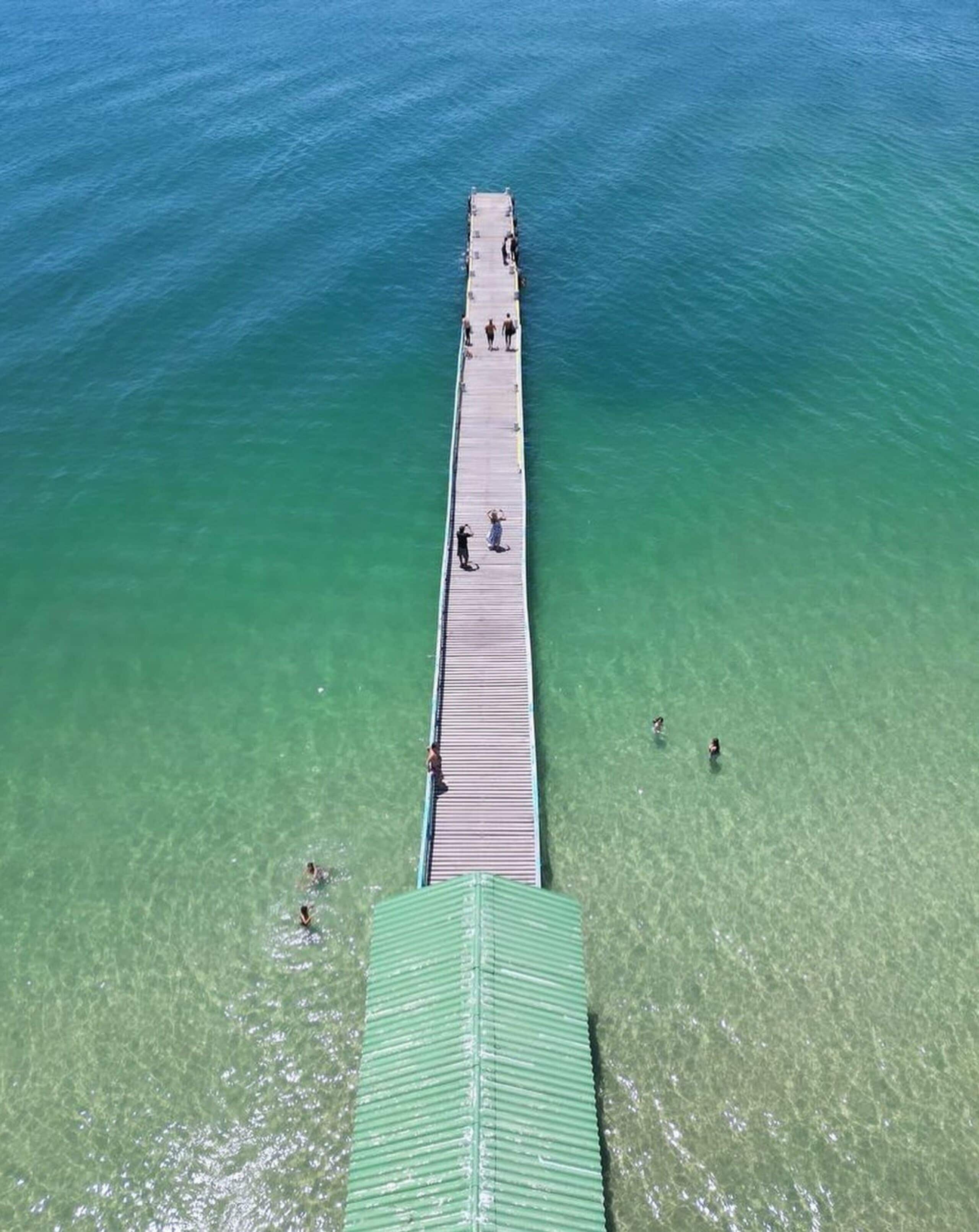 Vista aérea de un muelle que se adentra en aguas claras, con gente caminando por él.
