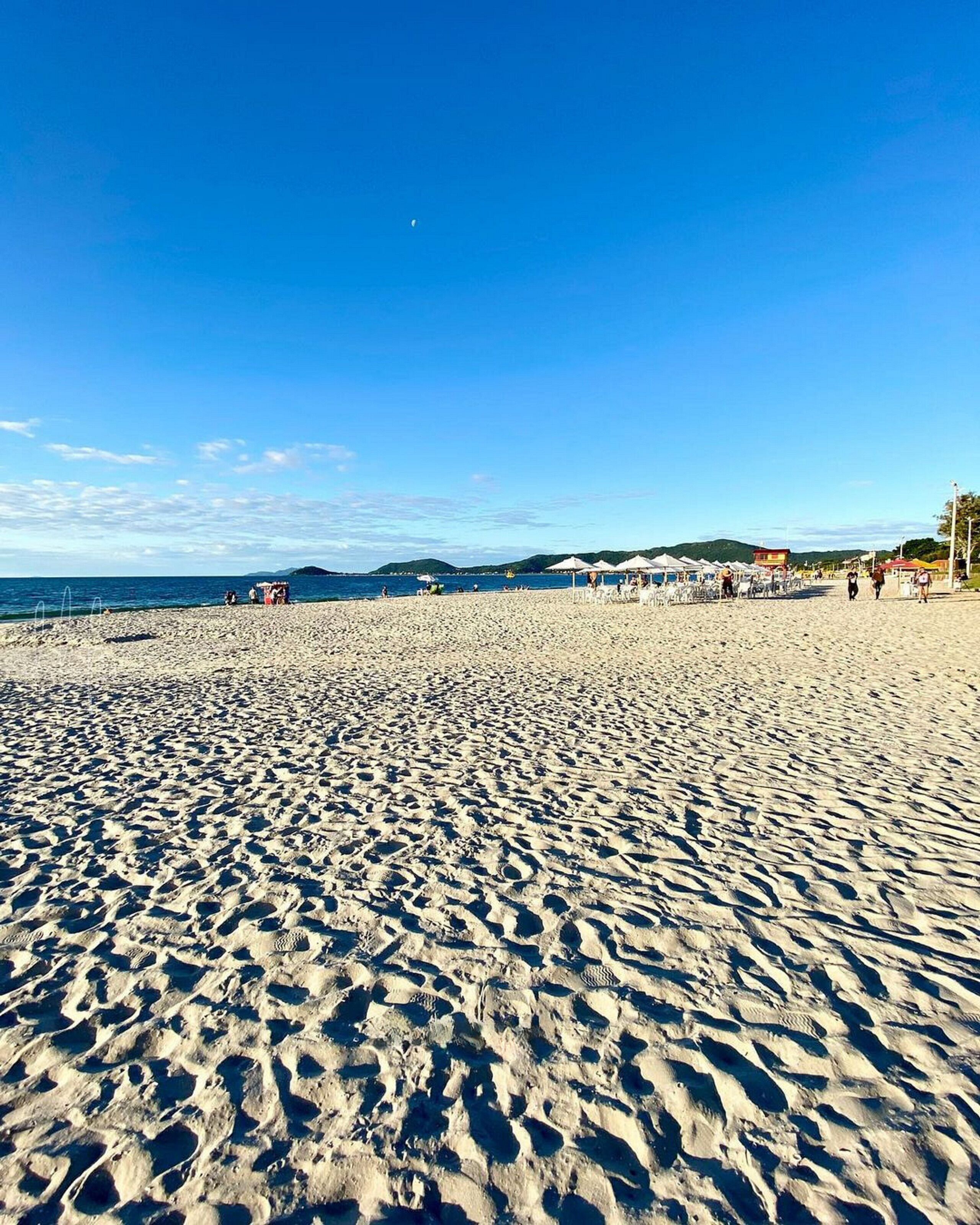 A wide sandy beach with umbrellas and beach chairs, a clear blue sky, and distant hills.