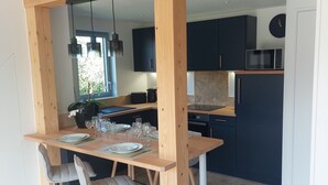Un coin repas avec une table et des chaises en bois, une cuisine à l’arrière-plan et une vue sur l’extérieur par une fenêtre.
