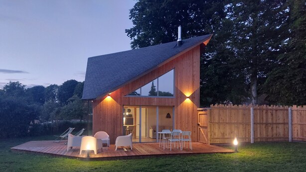 Une maison moderne dotée d’une terrasse en bois, d’un coin repas avec des chaises et d’une grande fenêtre.