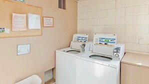 Une buanderie équipée de deux machines à laver à chargement frontal, d’un tableau d’affichage où sont affichées des avis et d’une poubelle.
