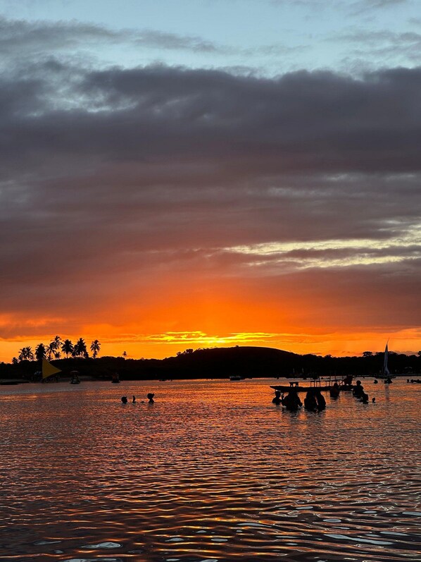 Atardecer sobre un cuerpo de agua con barcos y un velero, palmeras y una colina al fondo.