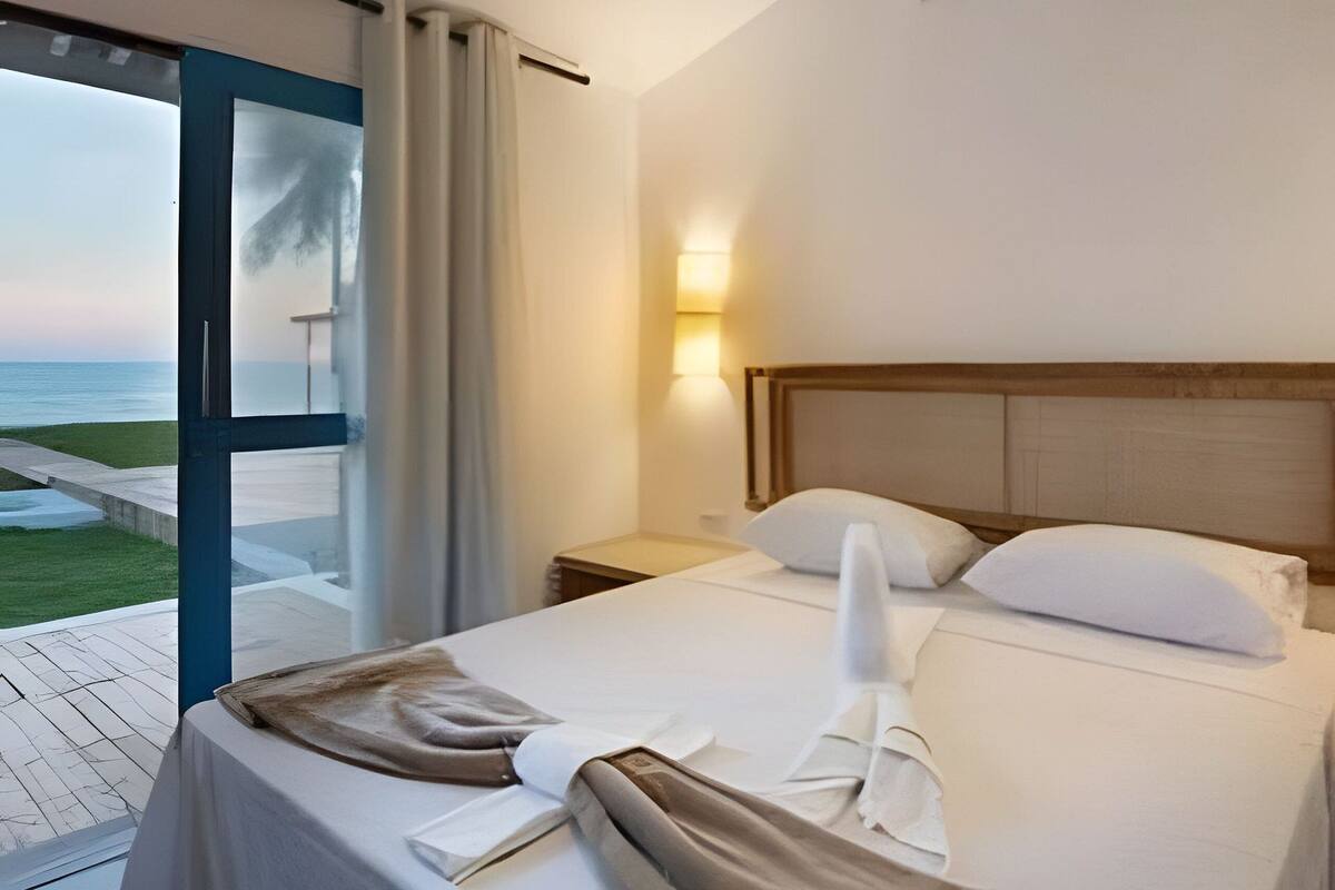 A hotel room with a bed, a nightstand, a lamp, and a view of the ocean through a glass door.