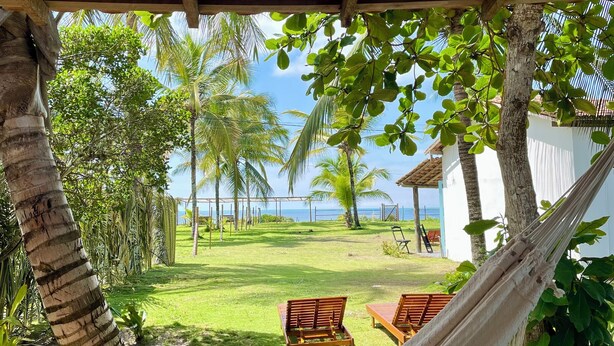 A hammock under a wooden pergola overlooking a grassy area with palm trees and a view of the sea. - Pousada Safyra  (Porto Seguro)