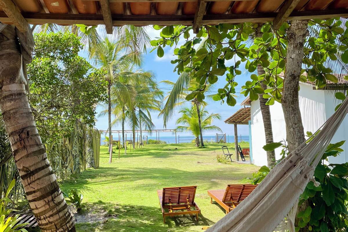 A hammock under a wooden pergola overlooking a grassy area with palm trees and a view of the sea.