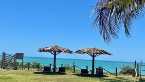 A beach view with two thatched umbrellas, a wooden fence, and a clear blue sky. - Pousada Safyra  (Porto Seguro)