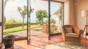 Una habitación con un gran ventanal corredero que da a un espacio exterior con una mesa y sillas, una silla de mimbre en el interior y una lámpara sobre una mesita.