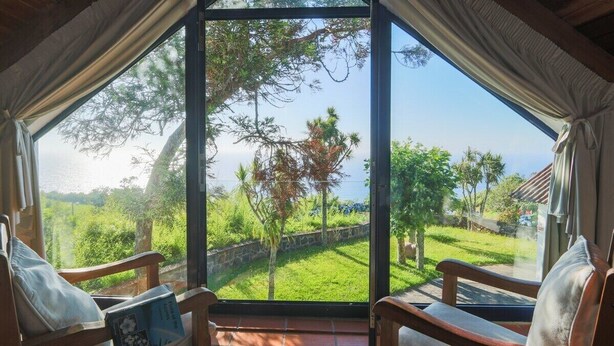 Una habitación con un ventanal amplio que ofrece vistas a un jardín exuberante y una extensión de agua. Hay dos sillas de madera con cojines.