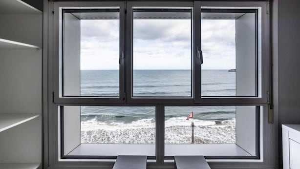 A room with a large window overlooking the ocean, a view of the beach, and a flag on the shore.
