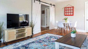 Una sala de estar con televisor, una puerta corrediza, una mesa de comedor con sillas y una ventana con marco rojo.