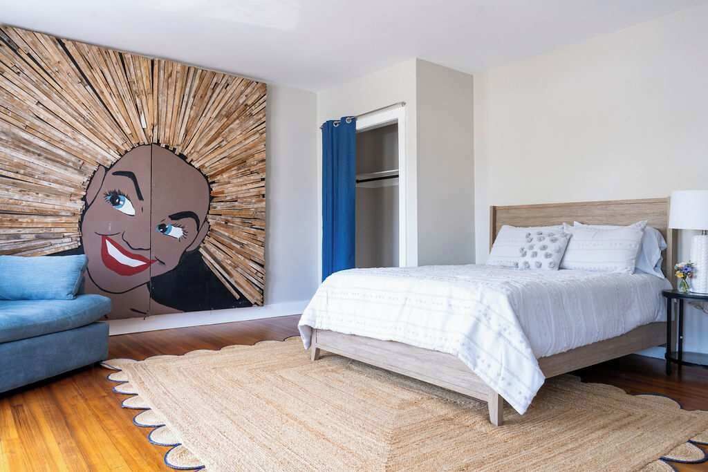 A bedroom with a bed, a blue chair, a wardrobe, and a wall with a large abstract design.