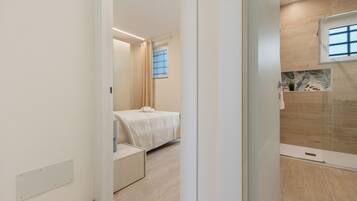 Ein modernes Hotelzimmer mit Bett, einer kleinen Sitzecke, einer Dusche und einem Fenster mit Jalousien.