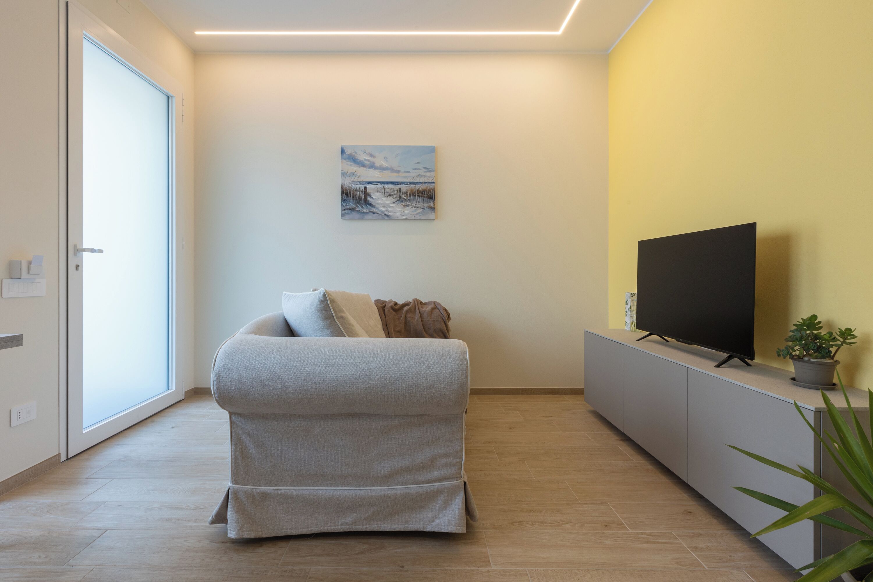 En moderne stue med en sofa, et fladskærms-tv på et stativ, et billede på væggen og et stort vindue.