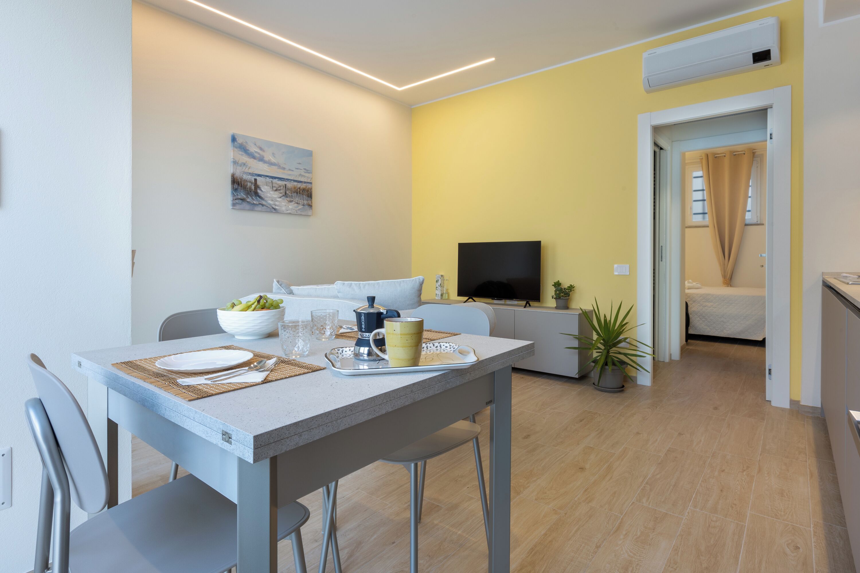 A modern living room with a dining area, a flat-screen TV, and a yellow accent wall.