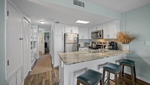 A modern kitchen with white cabinets, a stainless steel refrigerator, a microwave, and a marble island with bar stools.