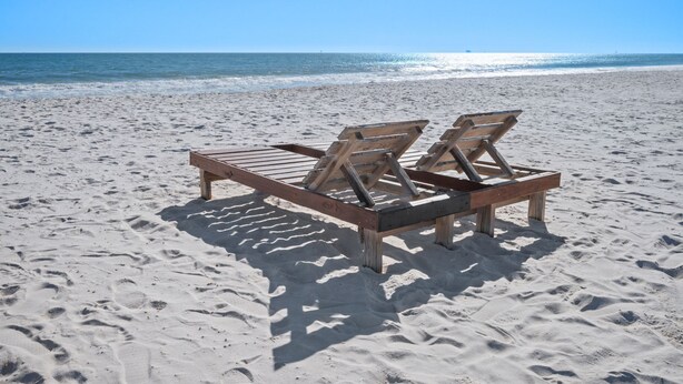 A wooden lounge chair on a sandy beach with footprints, a clear sky, and the ocean in the background.