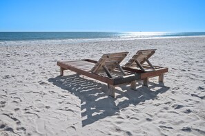 Una silla de madera en una playa arenosa con huellas, un cielo despejado y el océano al fondo.