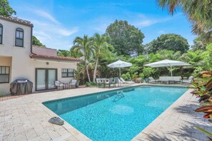 A swimming pool with lounge chairs and umbrellas, surrounded by palm trees and a building with a tiled roof. - Casita Atala - Nestle into a Tropical Designer Retreat with Shared Pool (West Palm Beach)