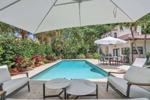 A swimming pool with lounge chairs and a table, surrounded by lush greenery and a white umbrella. - Casita Atala - Nestle into a Tropical Designer Retreat with Shared Pool (West Palm Beach)