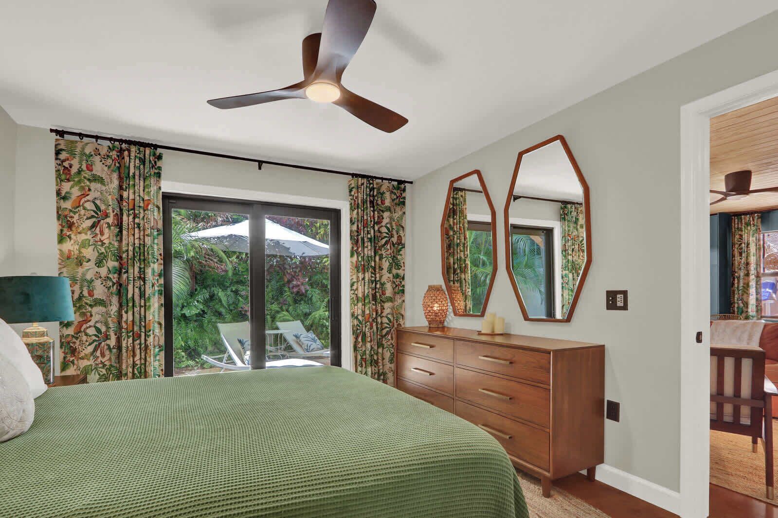 A bedroom with a bed, a dresser, a ceiling fan, and a view of a garden through a sliding door.