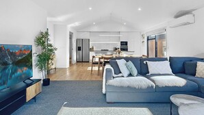 A modern living room with a sofa, a TV, and a dining area in the background. - Sunny Home Minutes to Remarkables Ski Field (Queenstown)