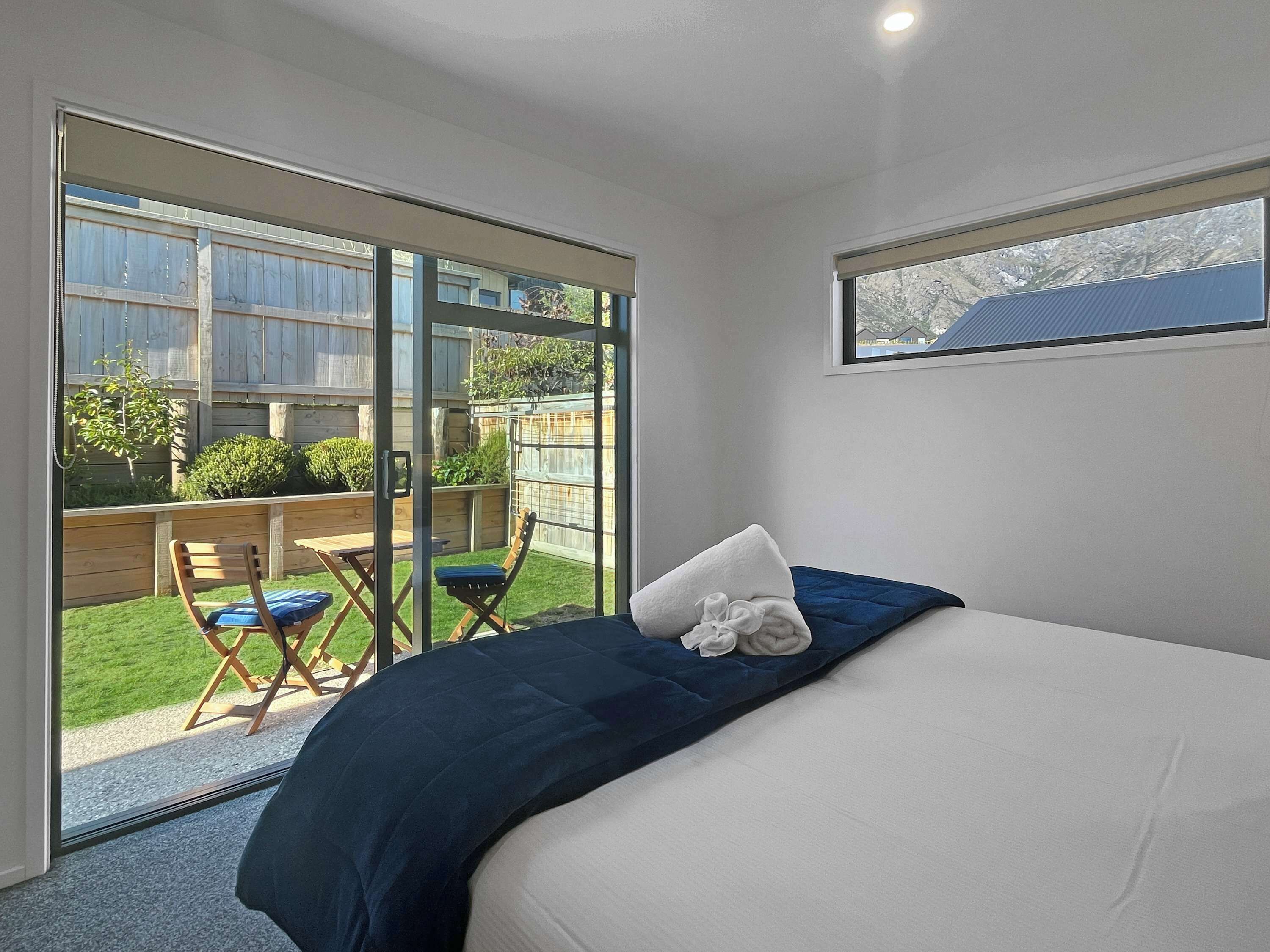 Ein Schlafzimmer mit Bett, Fenster und einer Schiebetür zu einer Terrasse mit Stuhl und kleinem Tisch.