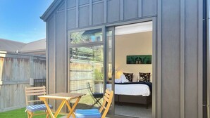 A small, modern building with a sliding glass door leading to a bedroom with a bed, pillows, and a painting. Outside, there is a patio with a table, two chairs, and a wooden deck. - Sunny Home Minutes to Remarkables Ski Field (Queenstown)
