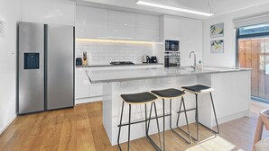 A modern kitchen with a stainless steel refrigerator, a built-in oven, and a gas stove. There are three black bar stools at the island. - Sunny Home Minutes to Remarkables Ski Field (Queenstown)