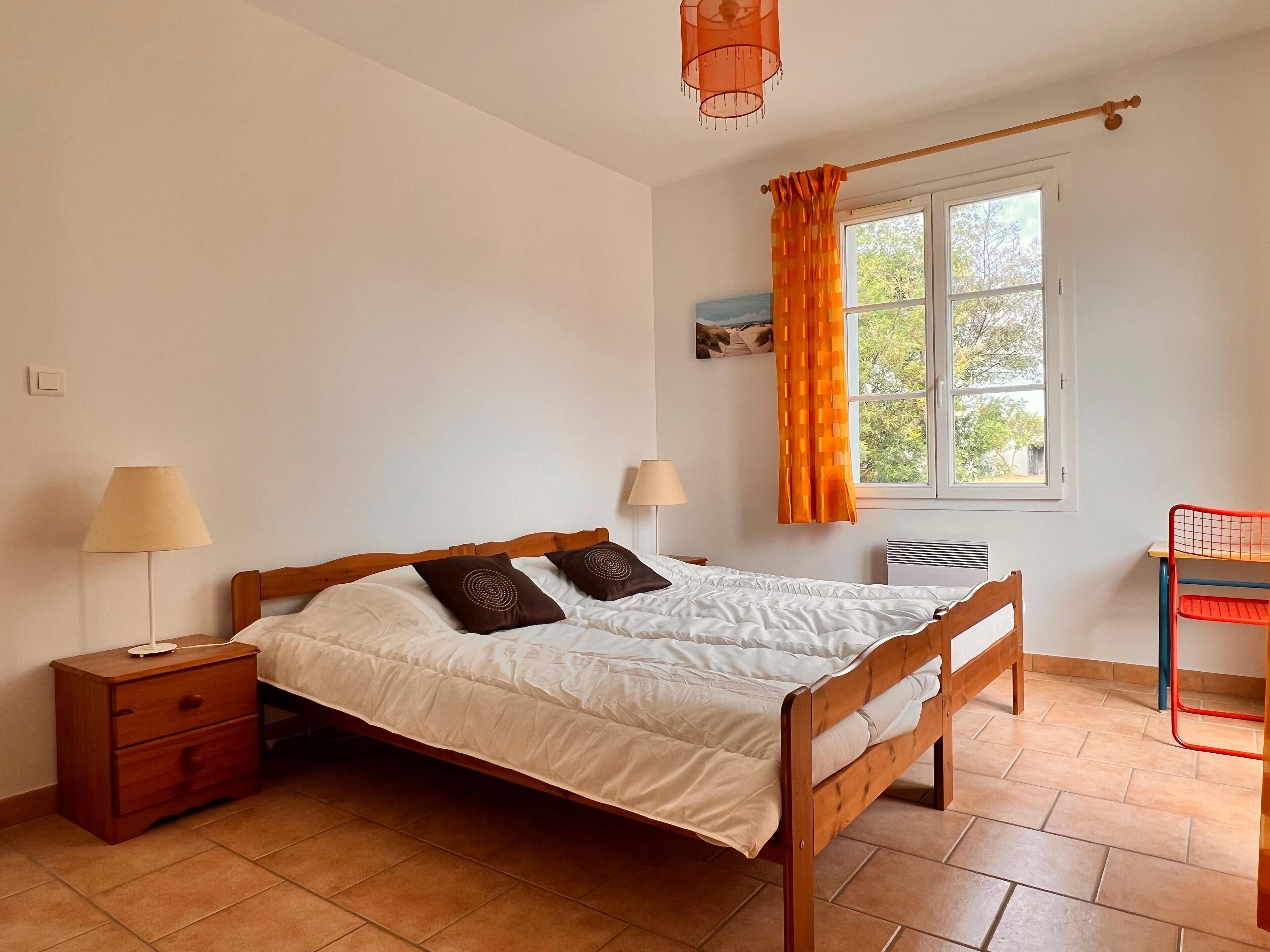 A bedroom with a wooden bed, two bedside tables, a window with orange curtains, and a red chair.