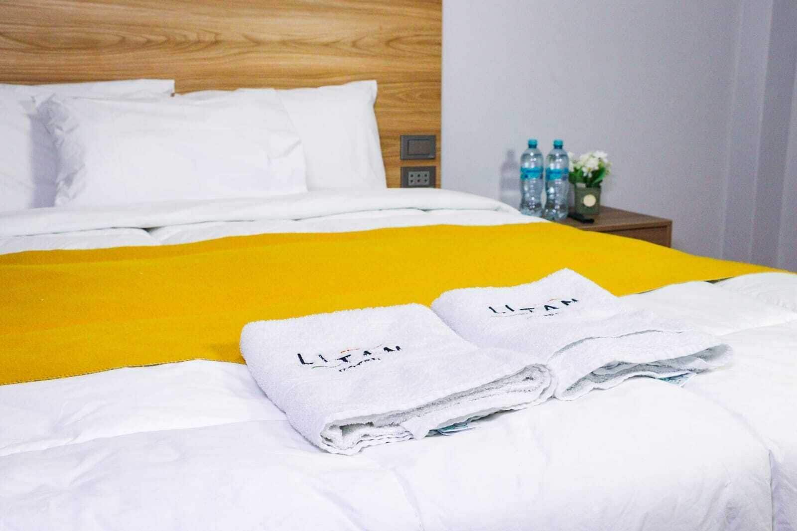 A neatly made bed with white linens and a yellow blanket, two folded towels with 'LITAM' on them, and a nightstand with water bottles and a vase of flowers.