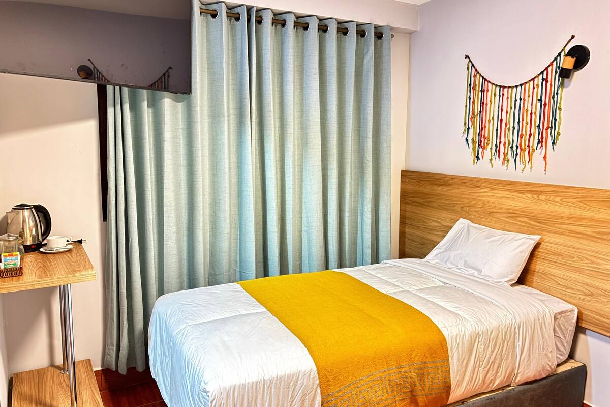 A hotel room with a bed, a wooden headboard, a wall-mounted light fixture, and a curtain.