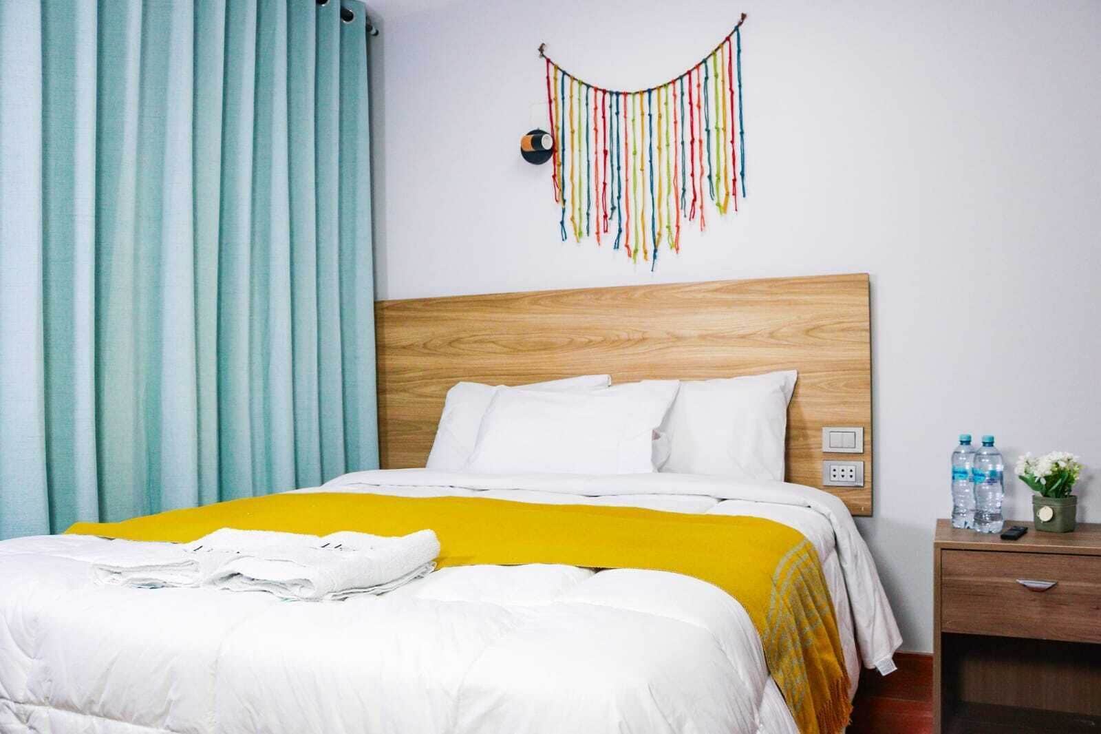 A hotel room with a wooden headboard, white bedding, a yellow throw blanket, a hanging decoration, and a nightstand with water bottles and a remote control.