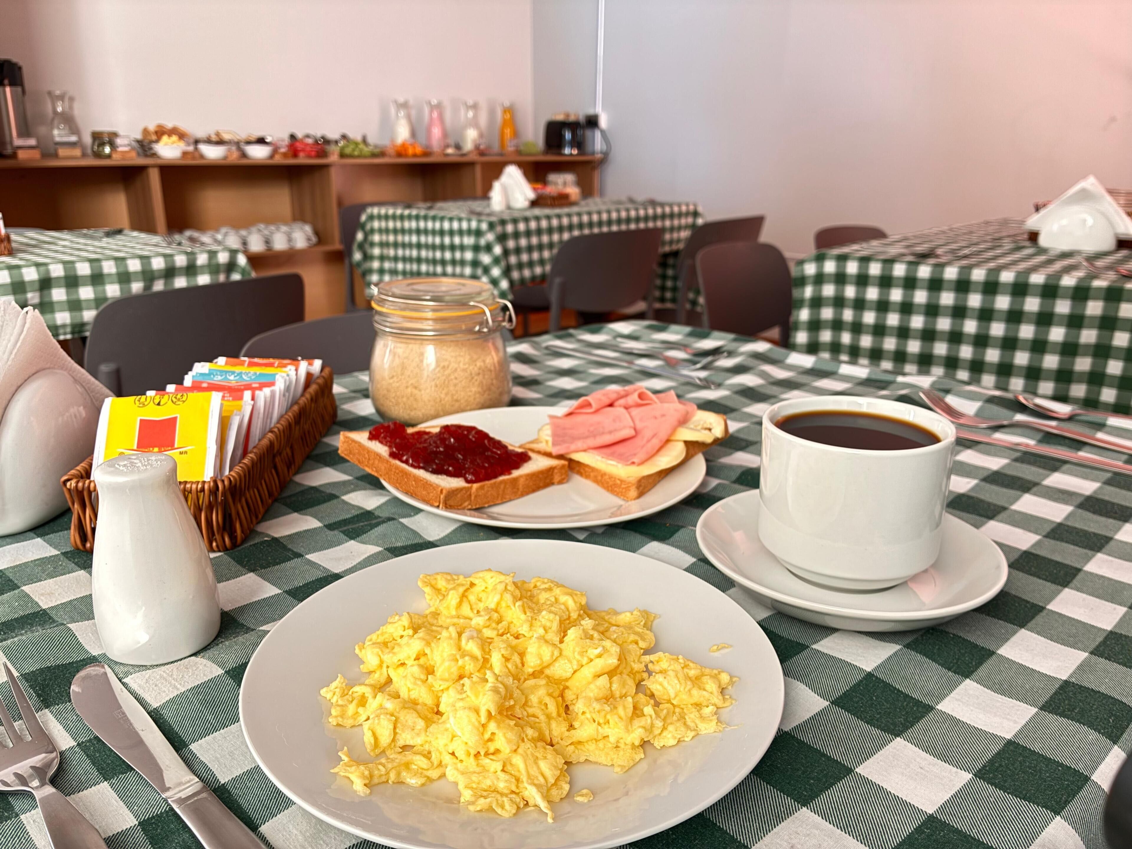 A breakfast setting with scrambled eggs, toast with jam and ham, coffee, and a milk jug on a checkered tablecloth.