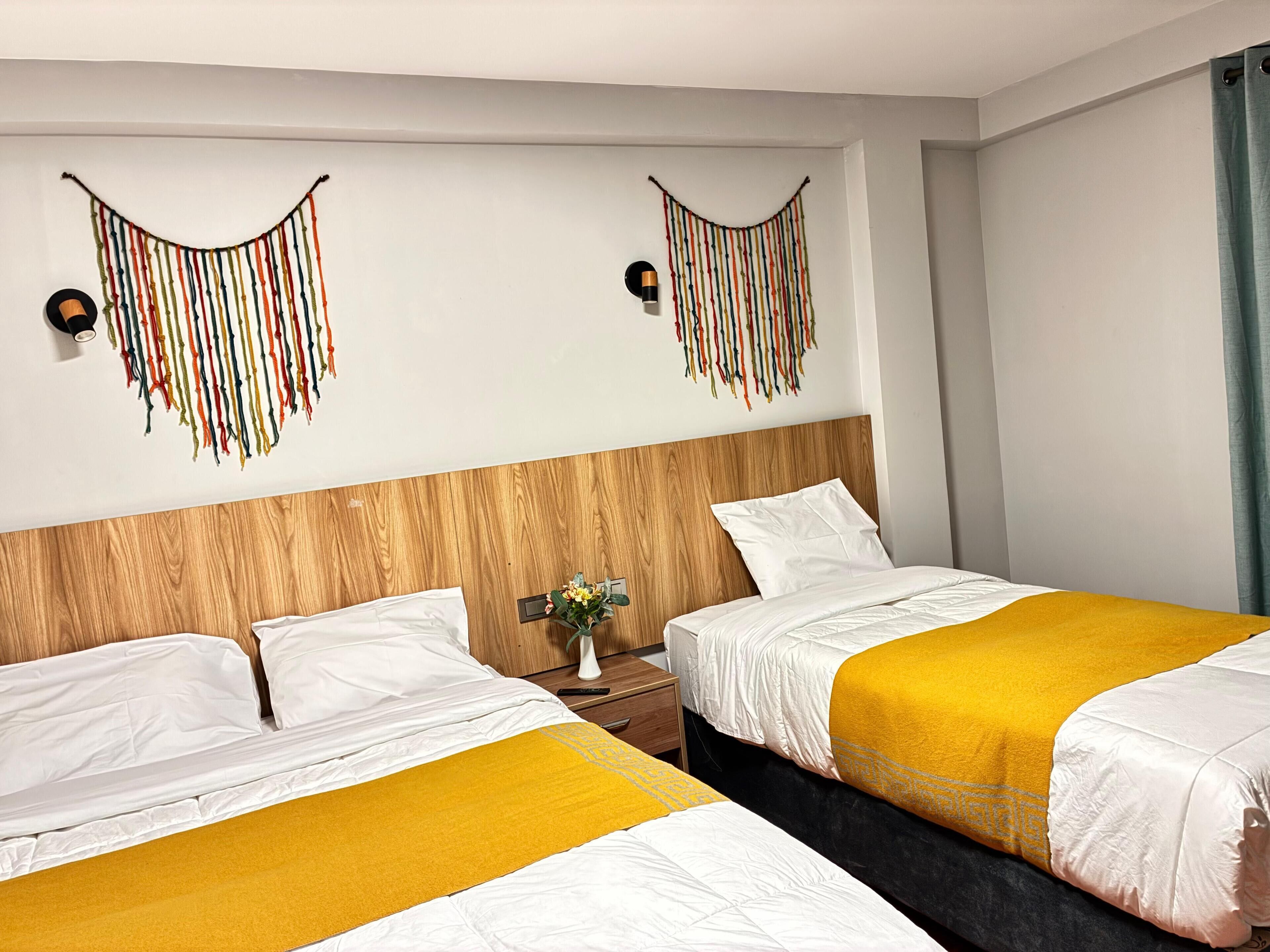 A hotel room with two beds, each with white bedding and a yellow accent. There are wall-mounted decorative items and a nightstand with a vase of flowers.