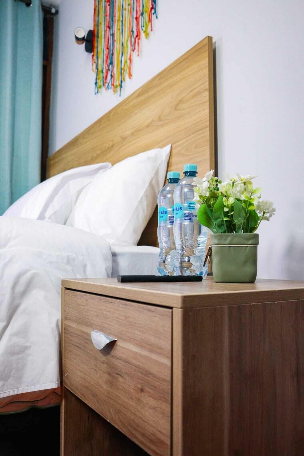 Un lit bien fait, avec du linge de lit blanc, une tête de lit en bois, une table de chevet ornée d’une plante verte et trois bouteilles d’eau.