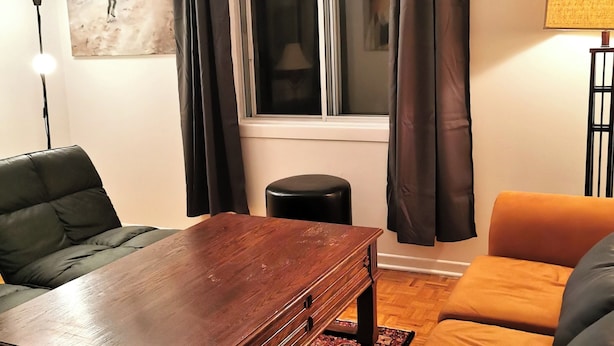 Ein Wohnzimmer mit einem Holztisch, einem orangefarbenen Sofa, einem schwarzen Ledersessel, einem schwarzen Hocker und einem Fenster mit Vorhängen.