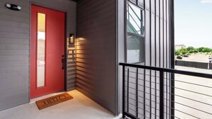 A red door with a doormat, a balcony with a metal railing, and a view of a parking lot.