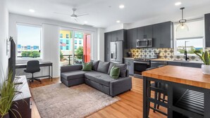 A modern apartment with a living area, kitchen, and dining space. It features a gray sectional sofa, a wooden dining table, and a kitchen with dark cabinets and stainless steel appliances.