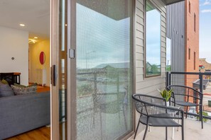 Een moderne woonkamer met een grijze bank, een ronde spiegel in het rood en een schuifdeur die leidt naar een balkon met zwarte stoelen en een plant in pot.