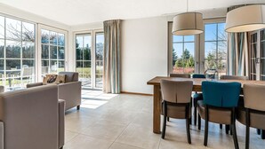Un salon spacieux avec une grande fenêtre, un coin repas avec une table et des chaises en bois, et un canapé agrémenté d’un coussin.
