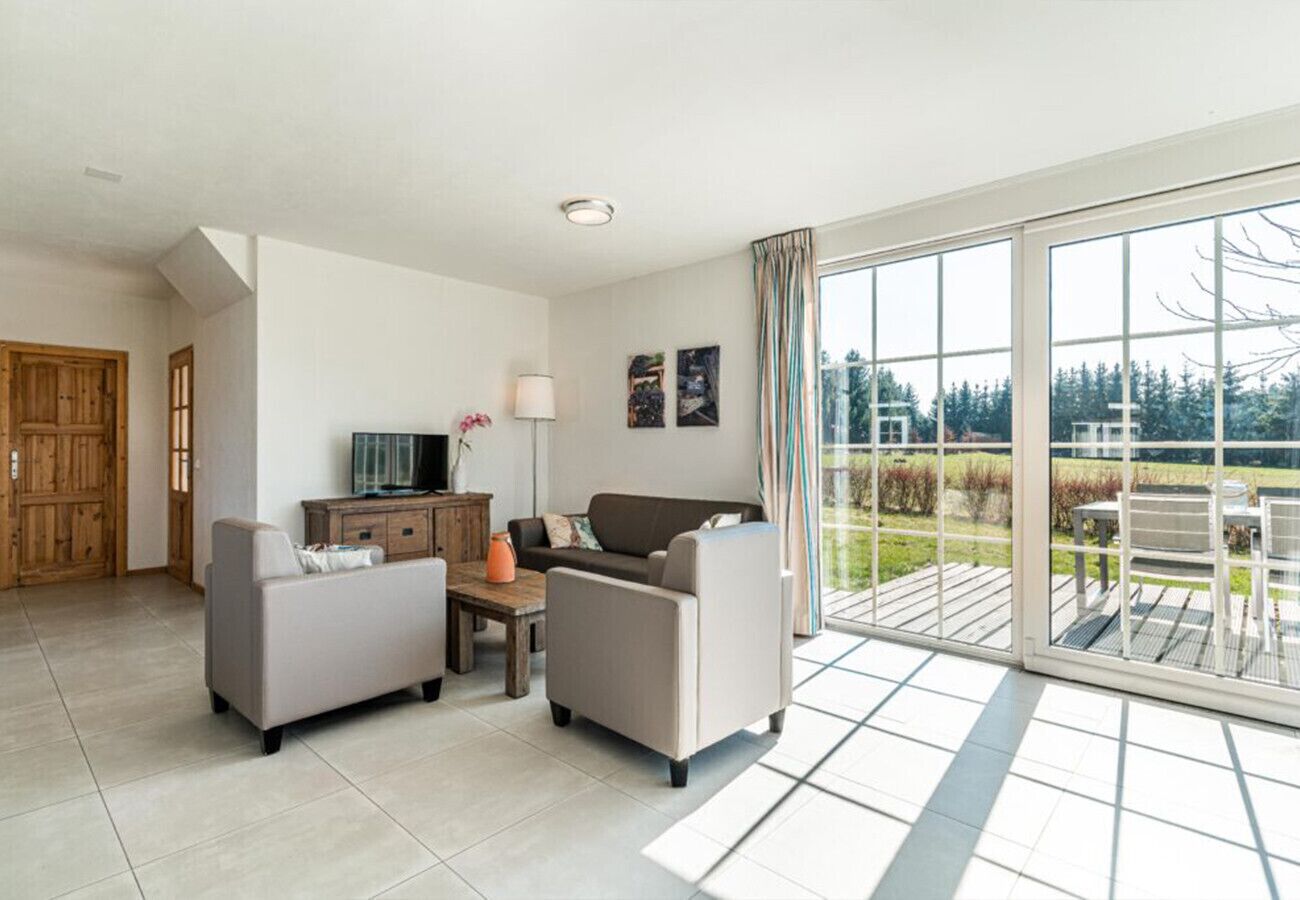 Una sala de estar moderna con un sofá, sillones, una mesa de centro y un televisor. Hay una puerta corrediza de vidrio que da a un balcón con mobiliario exterior.